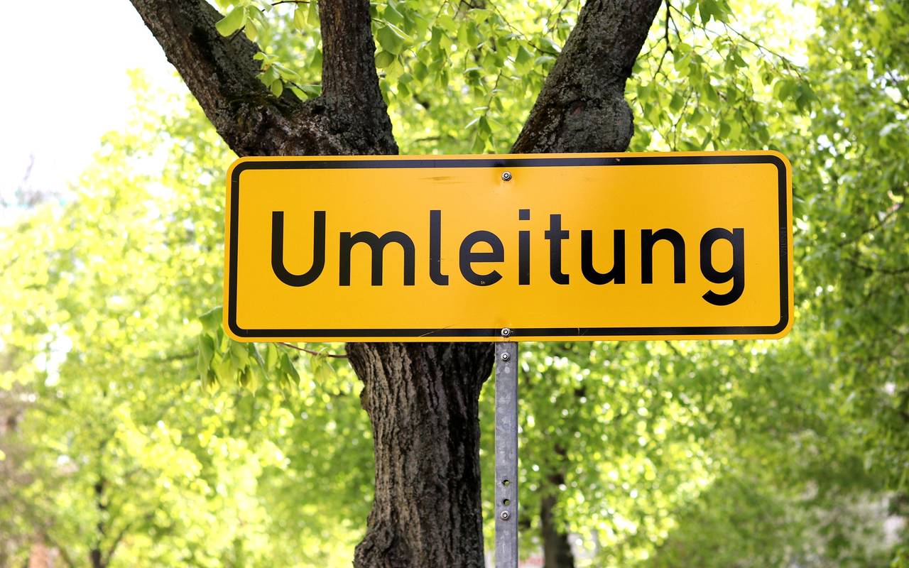 Ein Straßenschild aus Deutschland, das im Hintergrund gelb ist und die schwarze Aufschrift "Umleitung" trägt, steht vor einer Straßenkulisse mit einer begrünten Allee.